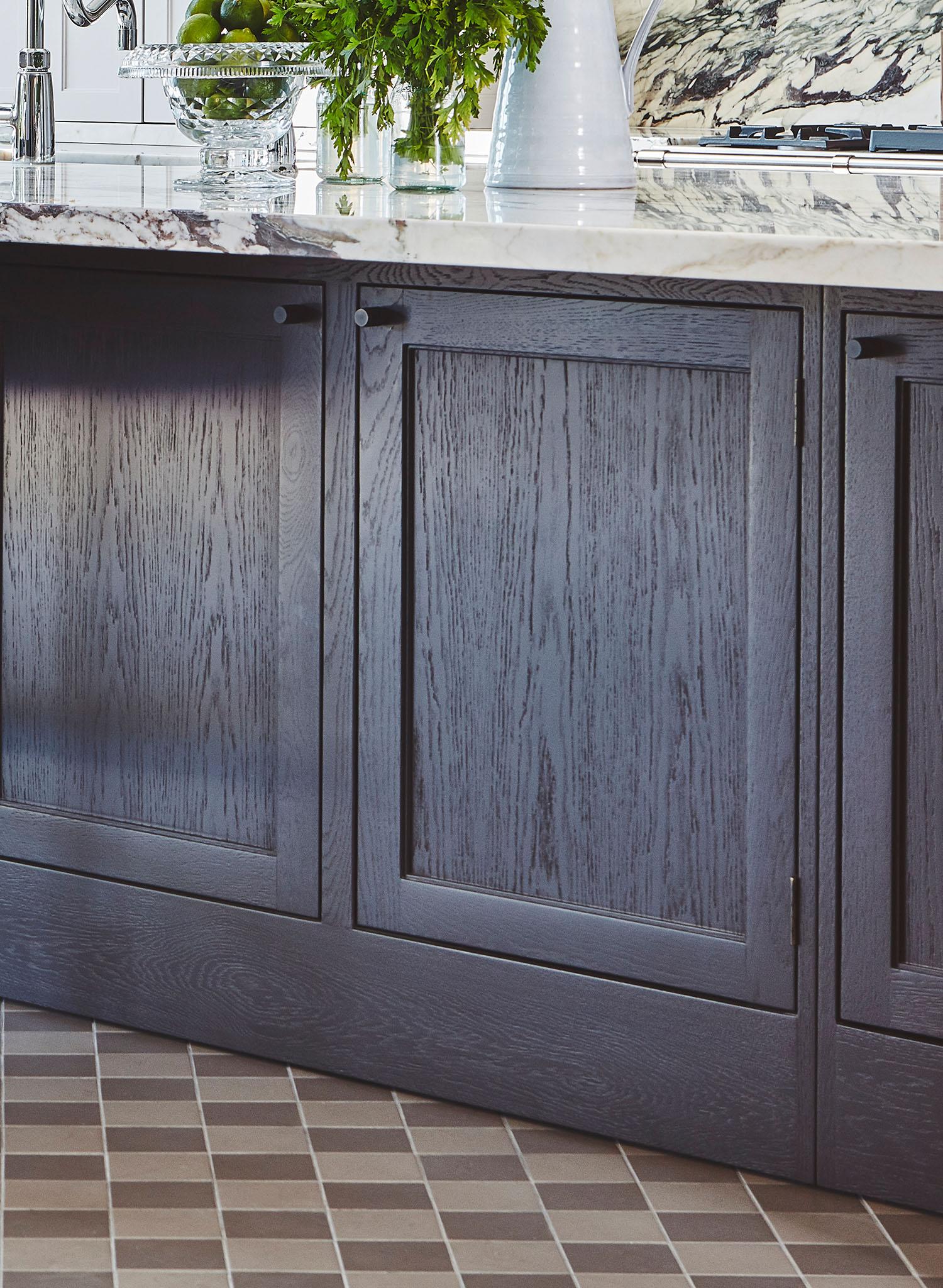 Dark wood kitchen cabinets with simple paneling, a marble countertop, a white vase with greenery, and a fruit bowl. The floor features brown and beige square tiles.