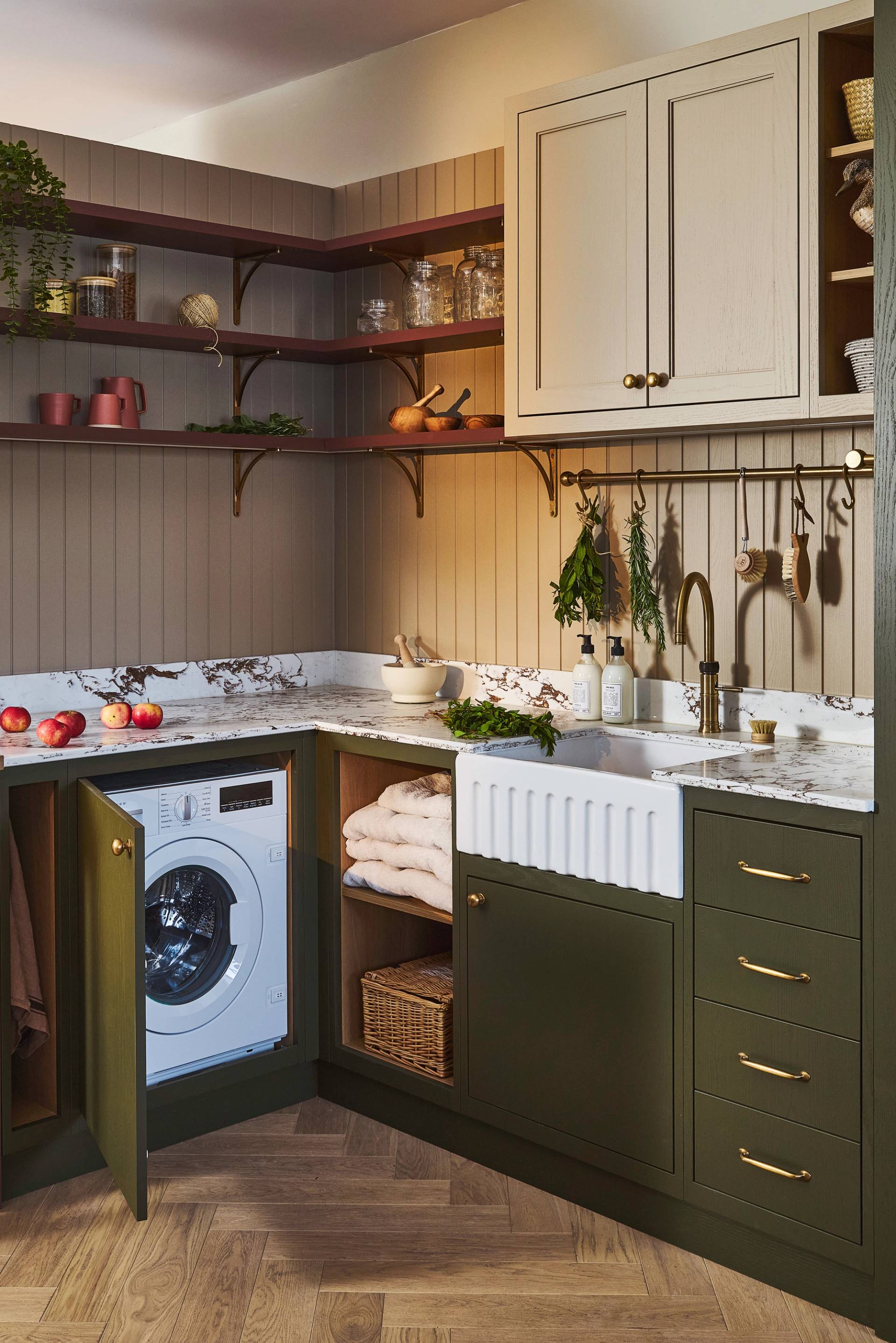 green cabinets in utility room with sink and cabinet open to reveal washing machine. There is a light coloured wall cabient and dark red coloured shelving. The shelves have pots and fresh herbs on them. The worktop is marble and is covered with red apples and more herbs