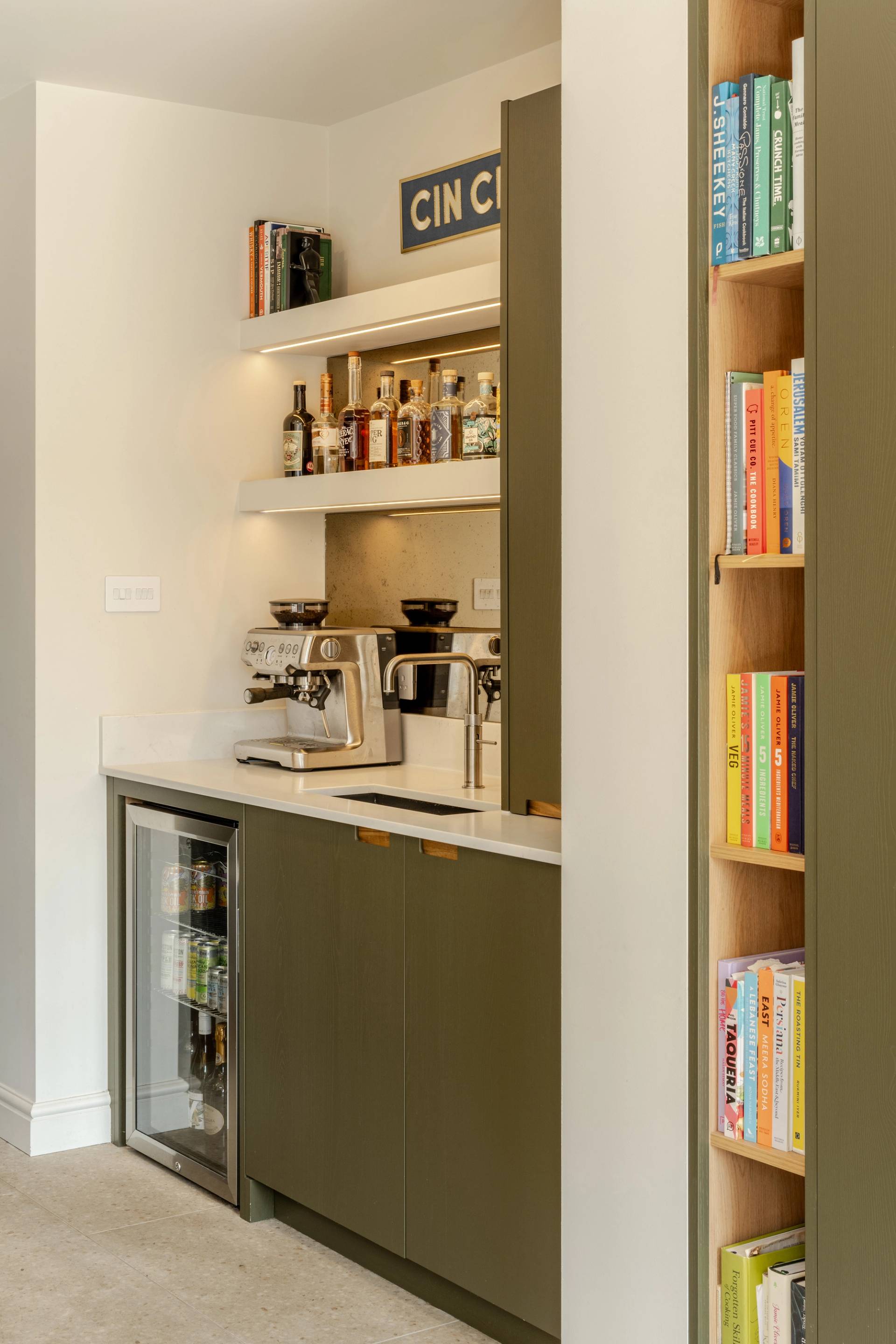 A modern kitchen with olive-green cabinets, open shelving with books and baskets, a small folding bicycle stored under a desk, and sunlight streaming through a glass door. A framed poster and potted plant are on the desk.