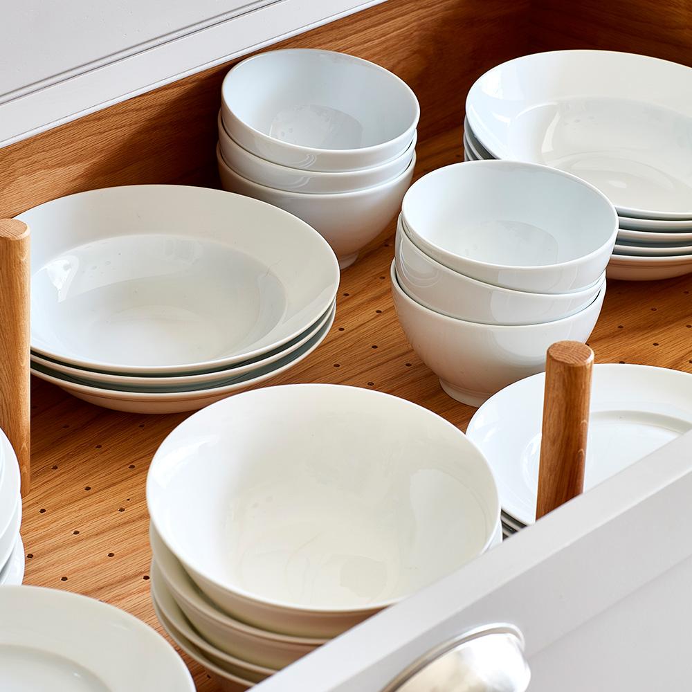 Open drawer with white ceramic bowls and plates neatly stacked on a wooden pegboard organizer, separated by wooden pegs.