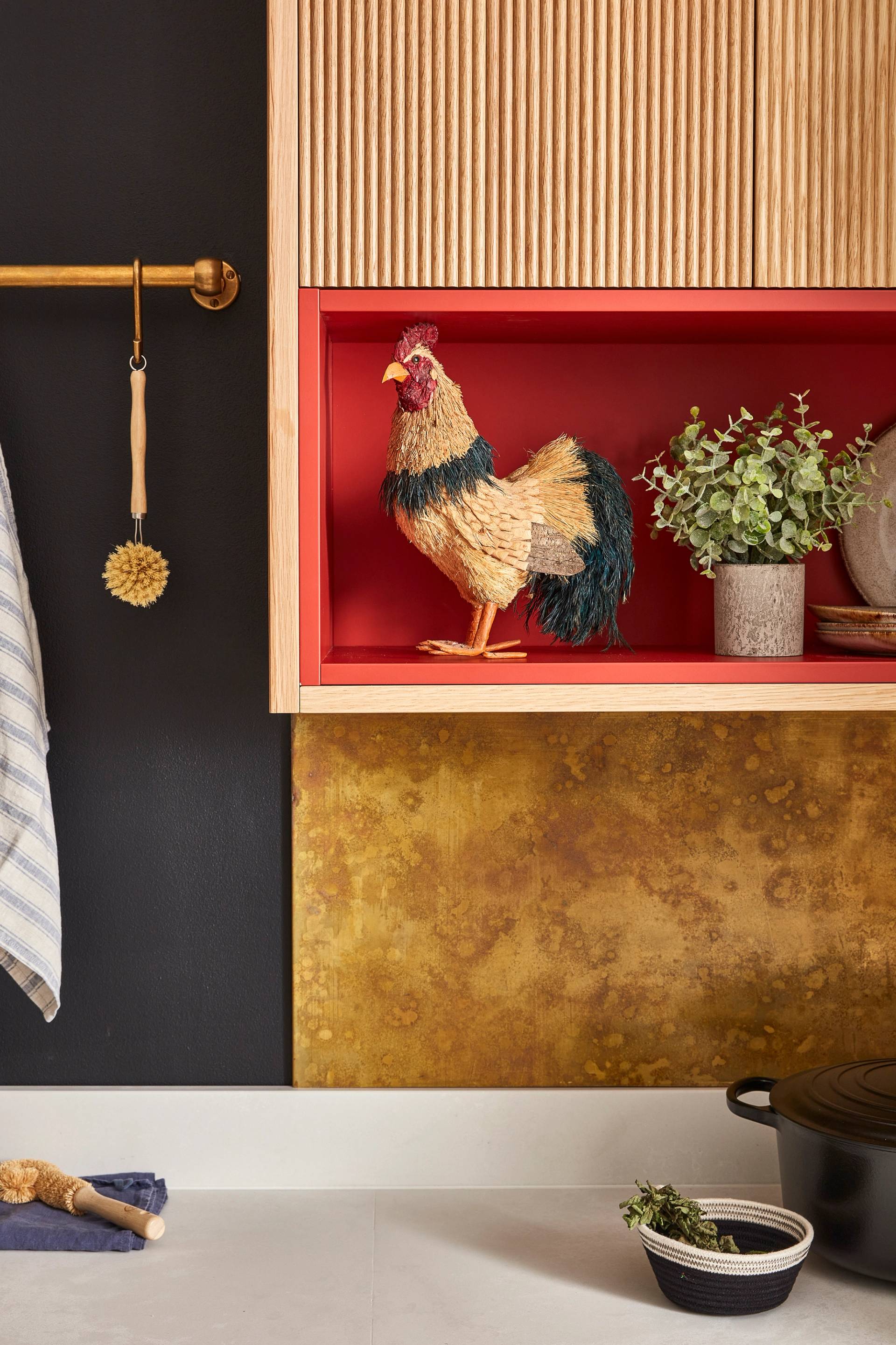 A kitchen with textured black and gold walls features a decorative rooster on a red shelf alongside green plants and stoneware. A gold rod holds a striped towel and brush. A black pot is on a white countertop below.