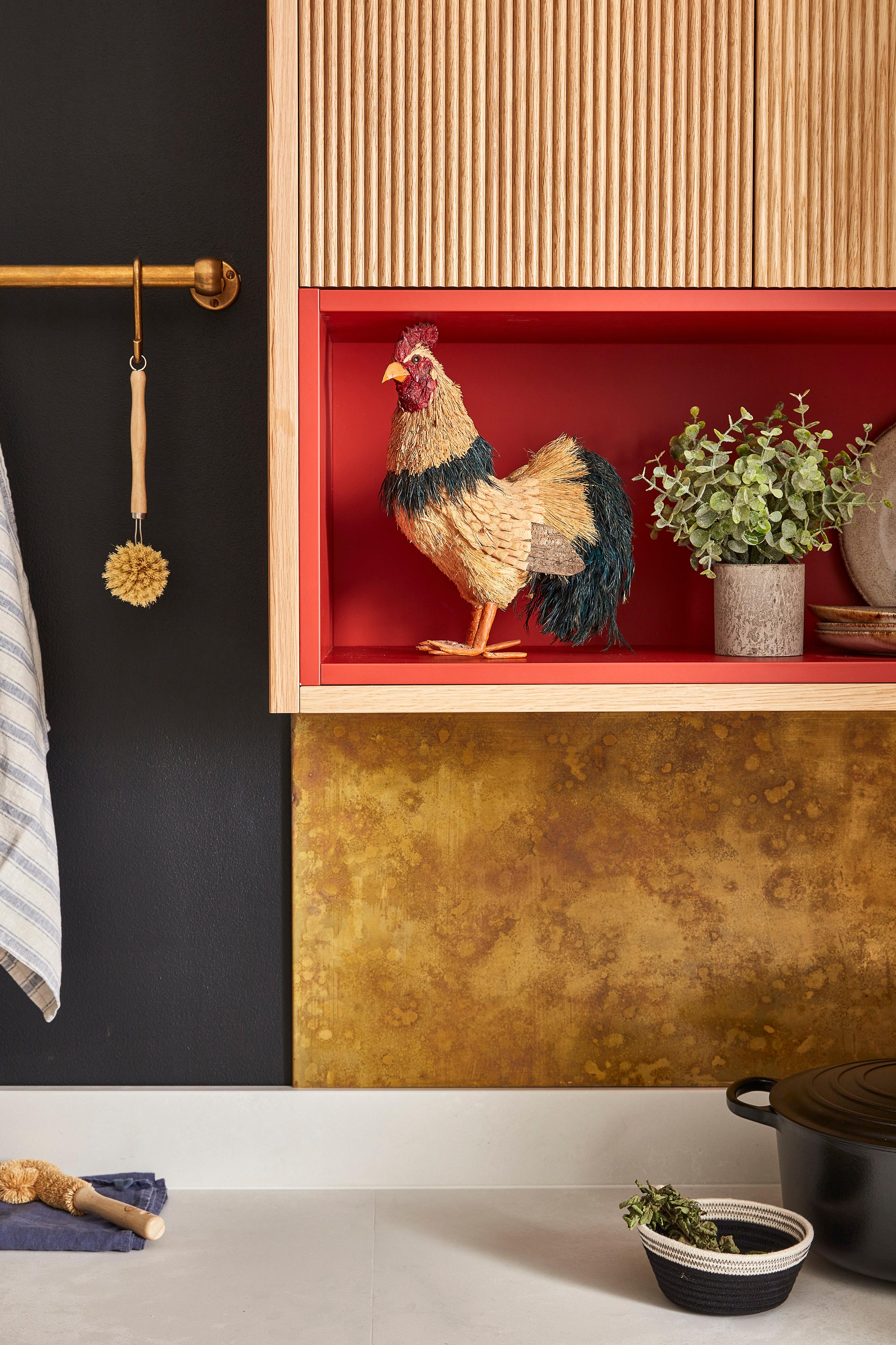 A kitchen with textured black and gold walls features a decorative rooster on a red shelf alongside green plants and stoneware. A gold rod holds a striped towel and brush. A black pot is on a white countertop below.