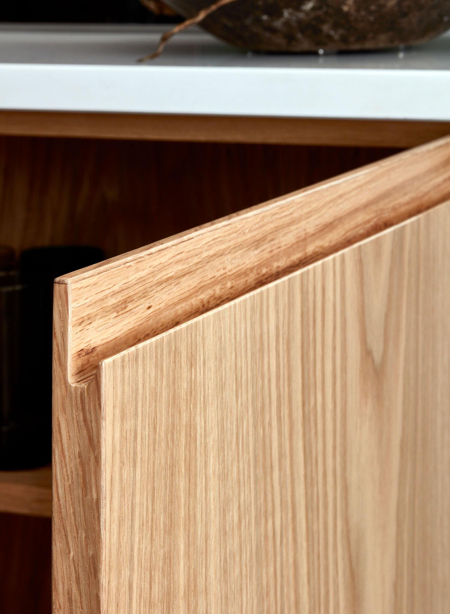 Close-up of a light wood kitchen cabinet door with a beveled edge, partially open to reveal part of the interior shelf. A dark object sits on the white countertop above the cabinet.