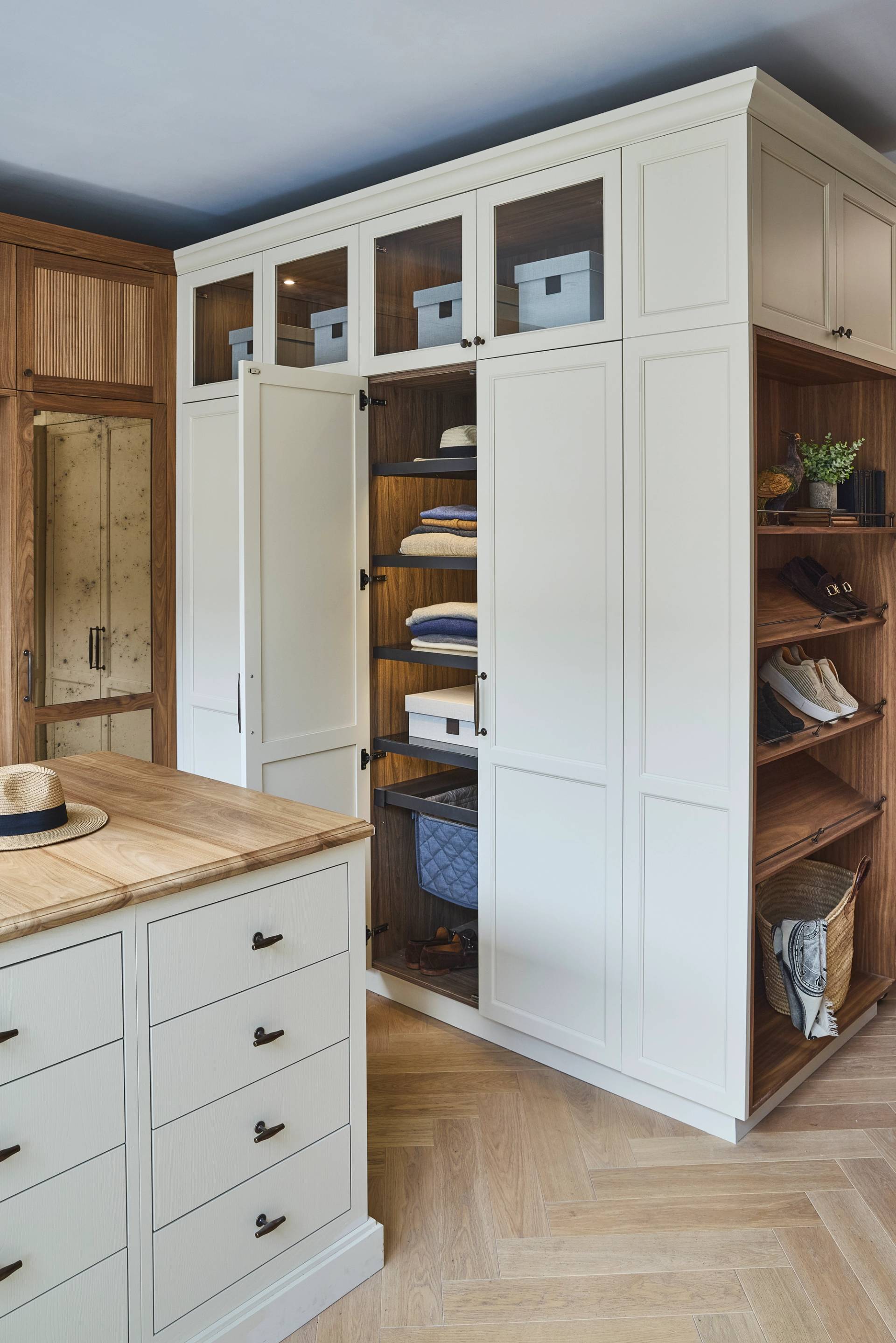 A modern walk-in closet with white and wood cabinetry, open shelves displaying neatly arranged hats, shoes, and bags, and an island with drawers on a light herringbone wood floor.