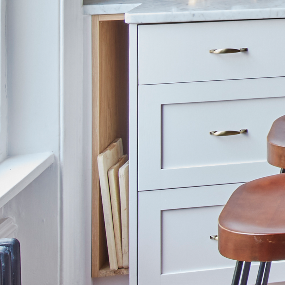 A close-up of a white kitchen cabinet with gold handles, a marble countertop, and a brown leather stool. A narrow vertical shelf next to the cabinet holds several cutting boards.