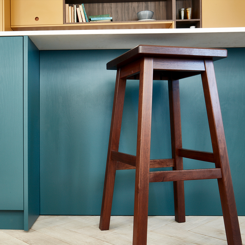 A tall wooden stool with a square seat stands in front of a blue kitchen island, with shelves and books visible in the background.