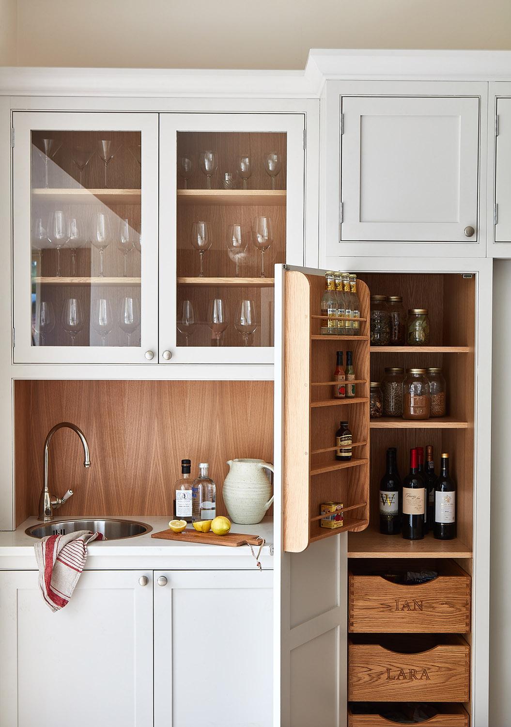 White kitchen cabinetry with glass display shelves for glassware, an open pantry showing wooden drawers and door storage, and a small bar sink area with warm wood paneling.
