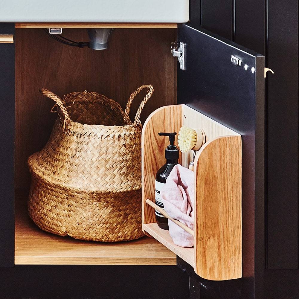 An open bathroom cabinet door with a wooden organizer holding a towel, soap dispenser, brush, and toothbrush. Inside the cabinet is a woven basket under the sink.