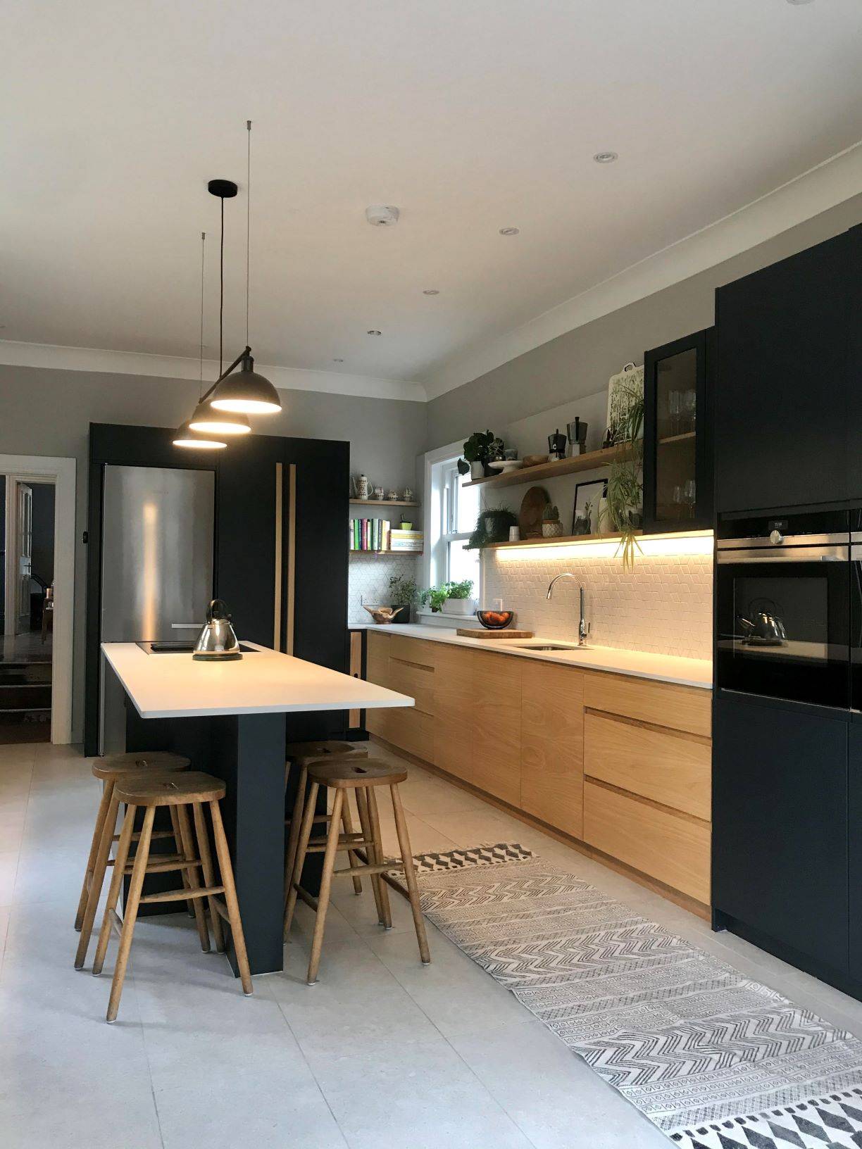 A modern kitchen with wooden cabinets and black accents. It features a central island with three wooden stools, pendant lighting, a stainless steel fridge, built-in oven, and open shelves with decor. The floor has a patterned rug.