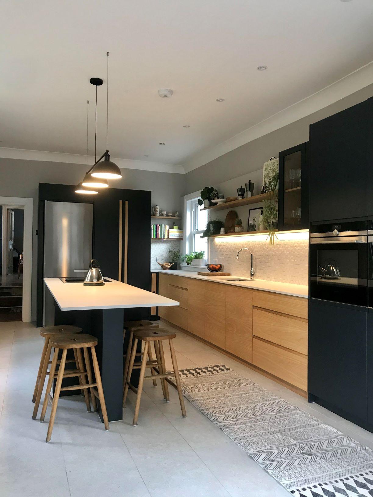 A modern kitchen with wooden cabinets and black accents. It features a central island with three wooden stools, pendant lighting, a stainless steel fridge, built-in oven, and open shelves with decor. The floor has a patterned rug.