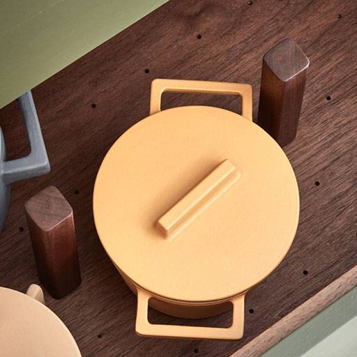 A yellow pot with a lid and rectangular handles is placed on a wooden surface next to dark wooden pegs. The scene is viewed from above, and part of another pot is visible in the corner.