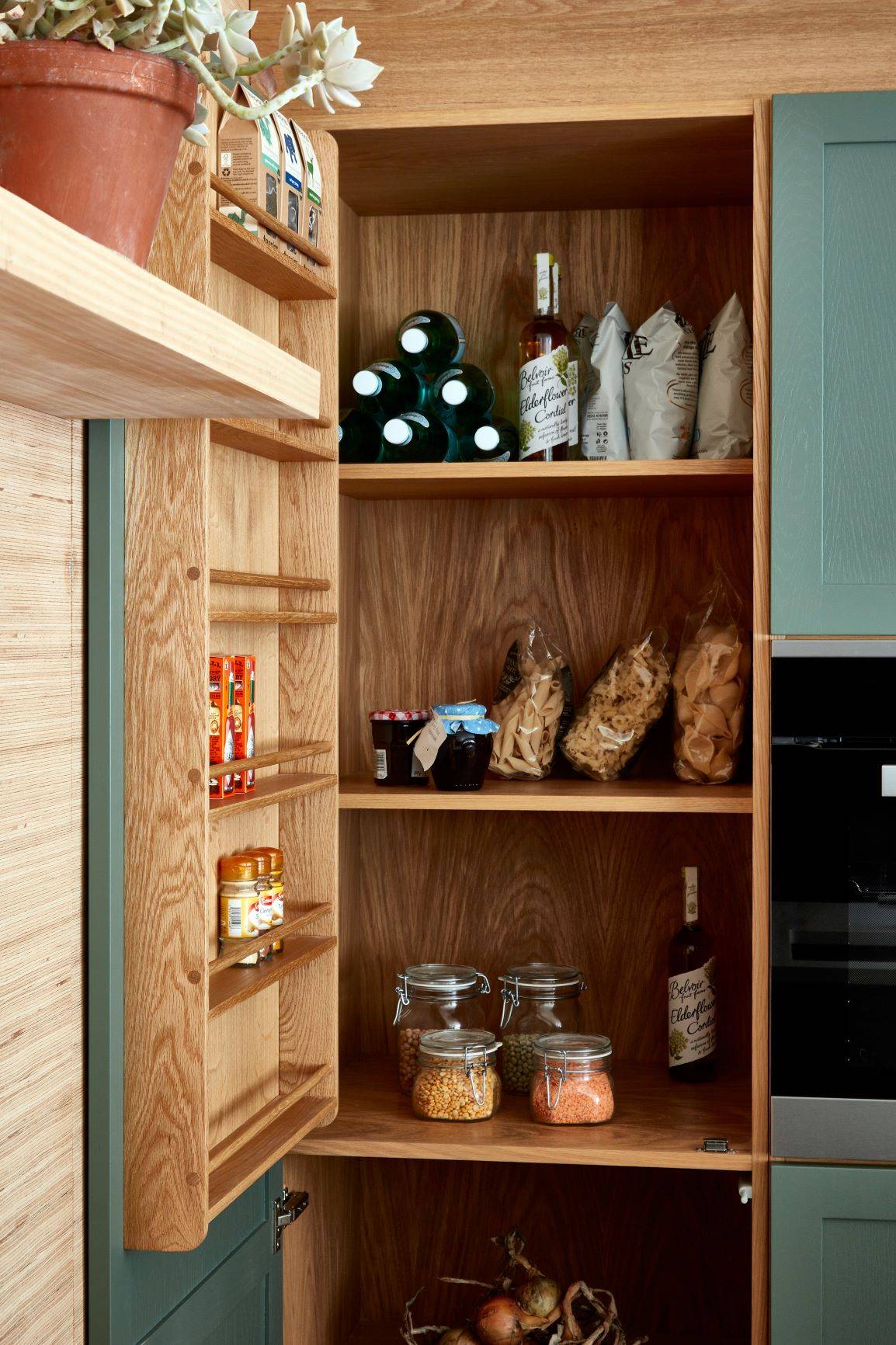 Open pantry with wooden shelves in a kitchen, holding various items including bottles of olive oil and wine, bags of chips, jars of pasta and grains, and canned goods. A potted plant sits on a nearby shelf. Blue cabinet doors are partially visible.