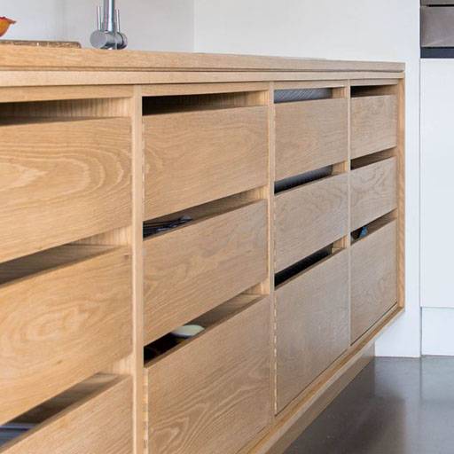 Light wood cabinets with modern, horizontal handles and a minimalist design. Some drawers are slightly open, revealing dishes inside. The cabinets are set against a light-colored wall and a polished floor.