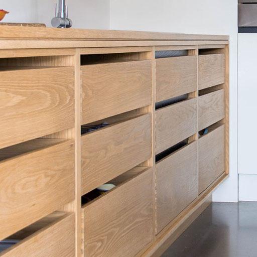 Light wood cabinets with modern, horizontal handles and a minimalist design. Some drawers are slightly open, revealing dishes inside. The cabinets are set against a light-colored wall and a polished floor.