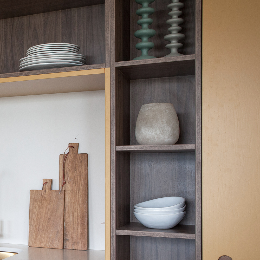 Open wooden shelving unit with stacked white plates, three large wooden cutting boards, three white bowls, a stone vase, and two green candlesticks arranged neatly on the shelves.