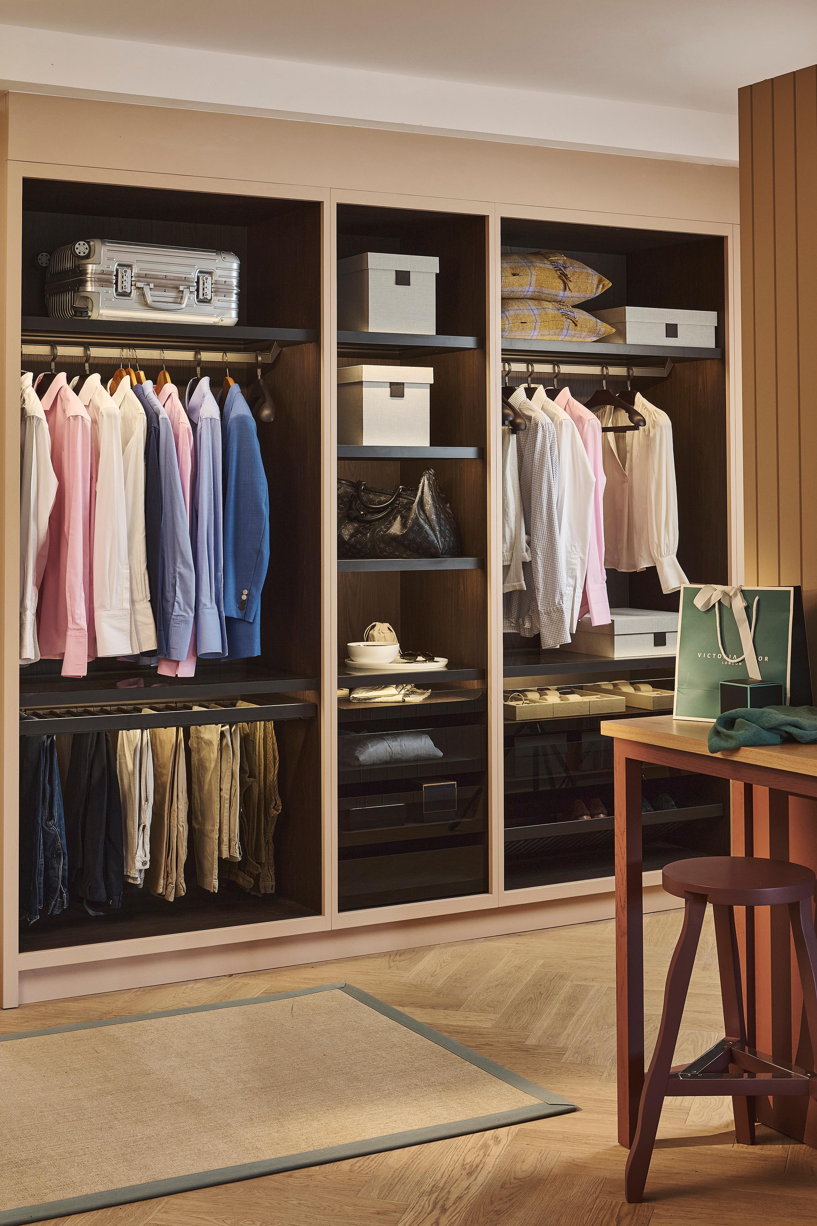 A neatly organized closet with shirts, jackets, and pants hanging, shelves with boxes and bags, folded clothes, and a suitcase. A wooden stool and shopping bag are visible in the foreground.