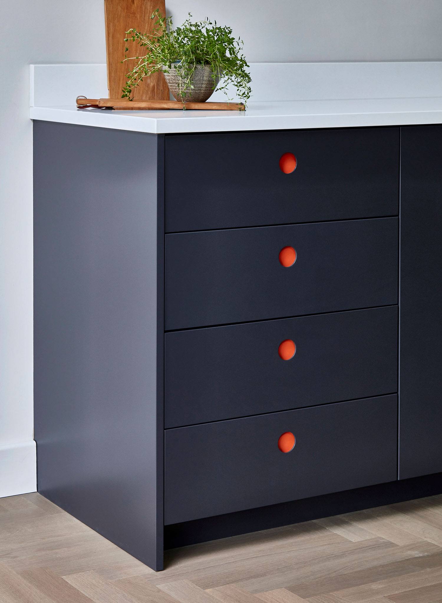 A modern kitchen cabinet with dark blue drawers, each featuring a round orange handle. On the white countertop above, there is a small potted plant and a wooden cutting board.