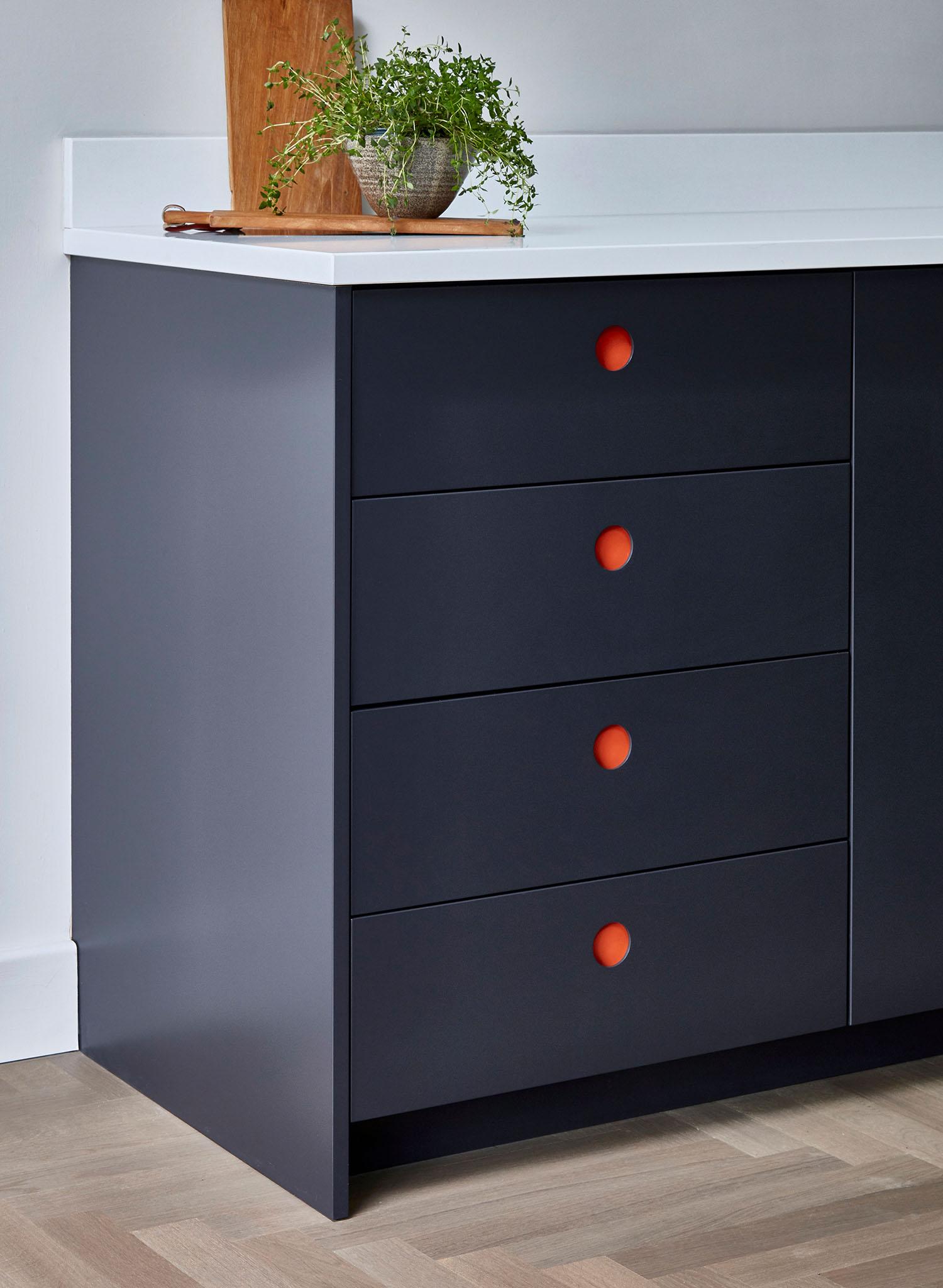 A modern kitchen cabinet with dark blue drawers, each featuring a round orange handle. On the white countertop above, there is a small potted plant and a wooden cutting board.