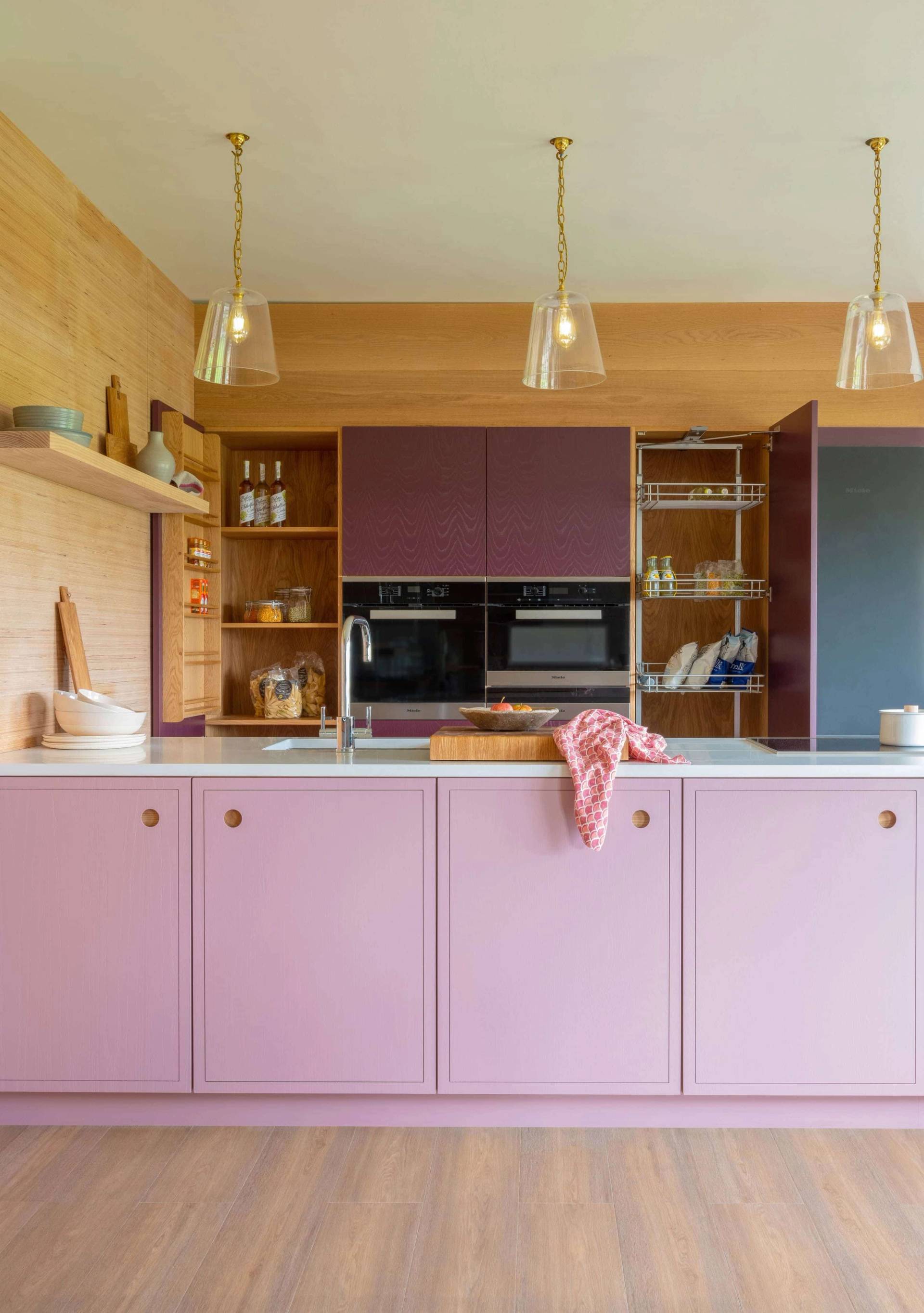 A modern kitchen with pink cabinets, wooden open shelves, and a light wood accent wall. Three pendant lights hang from the ceiling. A countertop features a cutting board with a pink cloth. Built-in appliances are visible in the background.