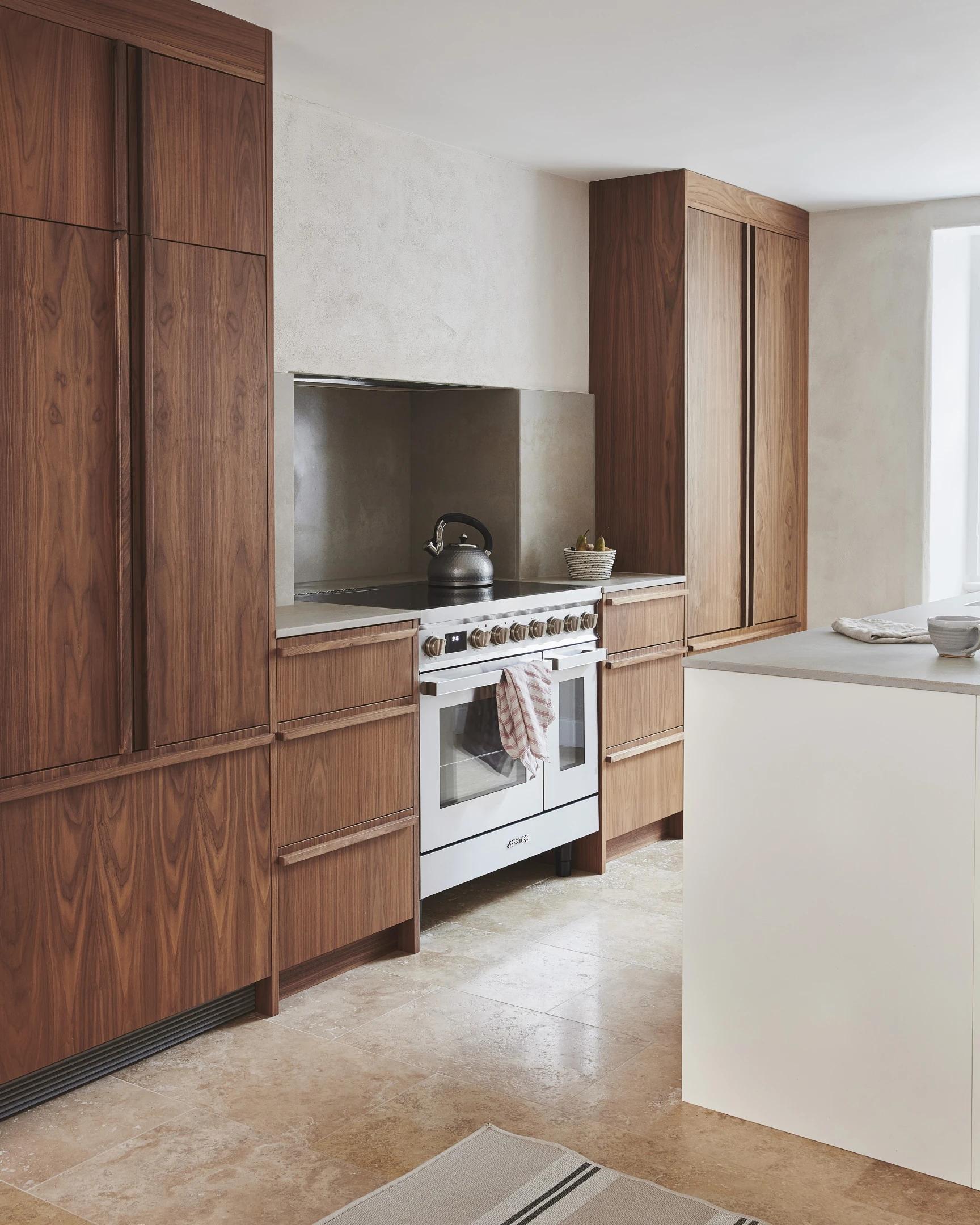 Modern kitchen with wooden cabinets, a white stove with a kettle on top, and a towel draped over the oven handle. A white countertop island is in the foreground, and a window allows natural light to fill the space.