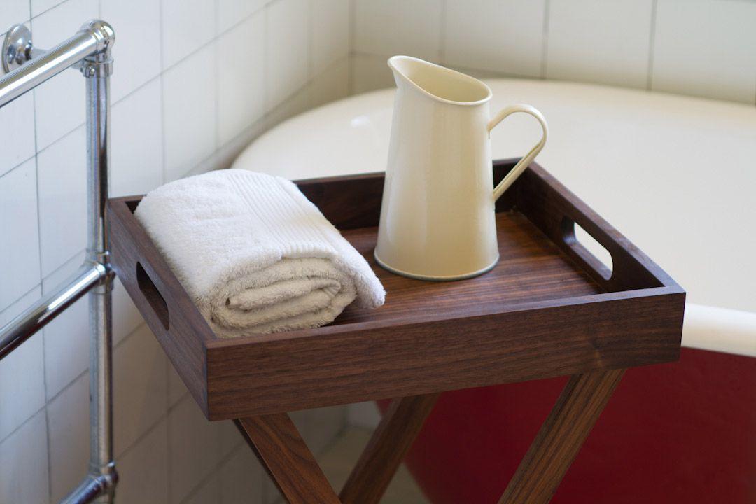 A wooden tray with a handle holds a folded white towel and a cream-colored pitcher. The tray is placed near a red bathtub against a wall with white tiles. Part of a metal towel rack is visible on the left.