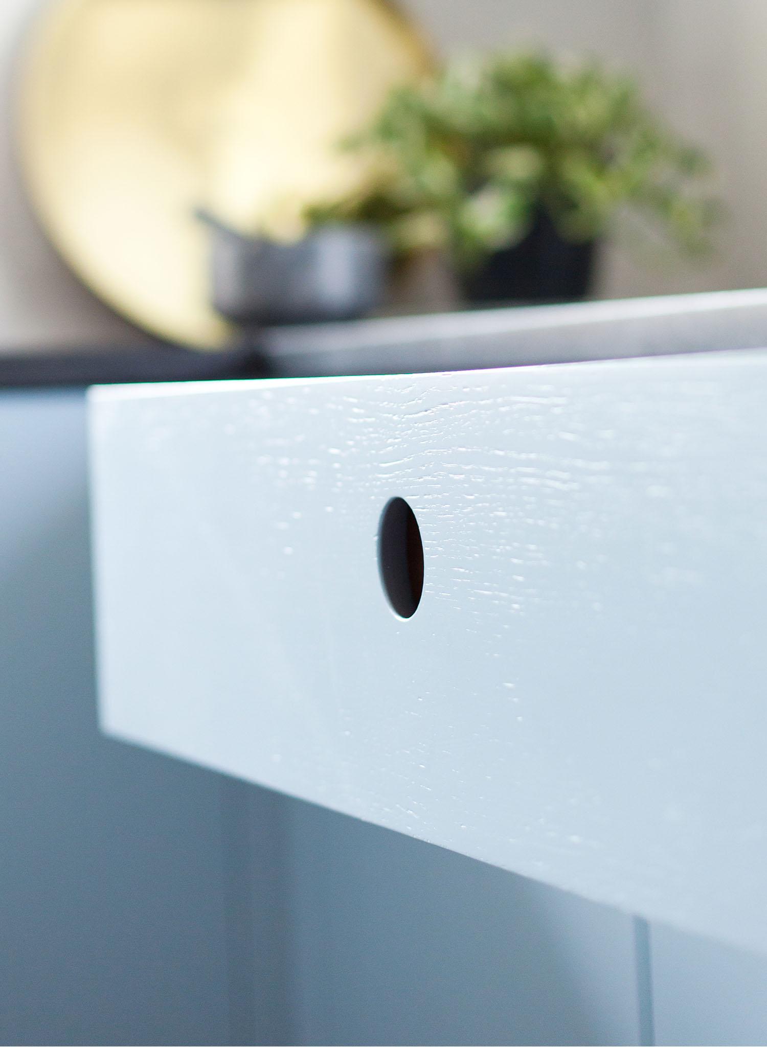 Close-up of a light blue drawer with a round cut-out handle, part of a cabinet. In the blurred background, there is a gold tray and a potted plant on a dark countertop