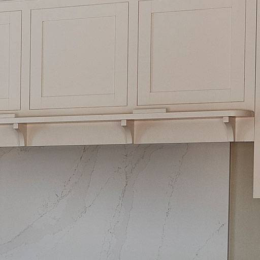 Cream-colored kitchen cabinets above a light marble backsplash with subtle gray veining. The cabinets have simple, recessed panel doors and decorative corbels supporting a small shelf beneath them.