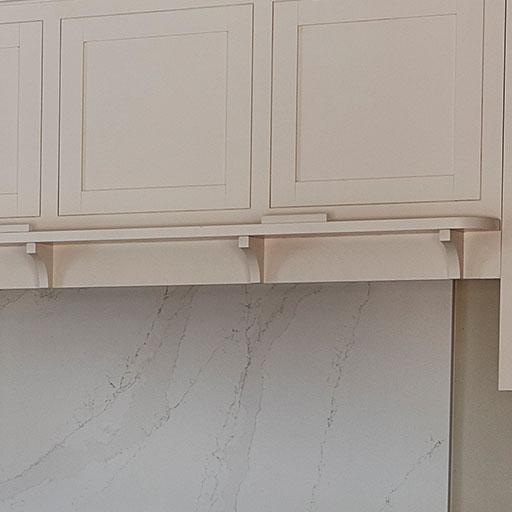 Cream-colored kitchen cabinets above a light marble backsplash with subtle gray veining. The cabinets have simple, recessed panel doors and decorative corbels supporting a small shelf beneath them.