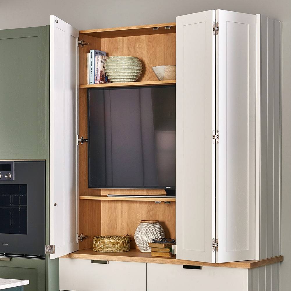 Kitchen cabinet with white doors open, revealing a television on a wooden shelf. Two green decorative bowls and a basket are on the shelves. The cabinet is integrated into a kitchen wall, next to a green oven unit.