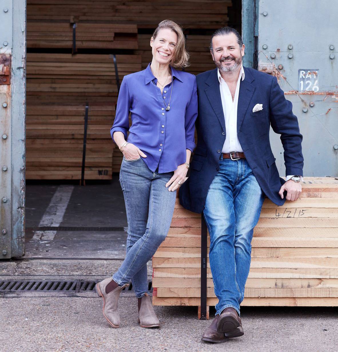 A woman and a man are leaning against a stack of wooden boards in front of a large open warehouse door. The woman is wearing a blue blouse and jeans, and the man is in a white shirt, blue blazer, and jeans. They are both smiling.