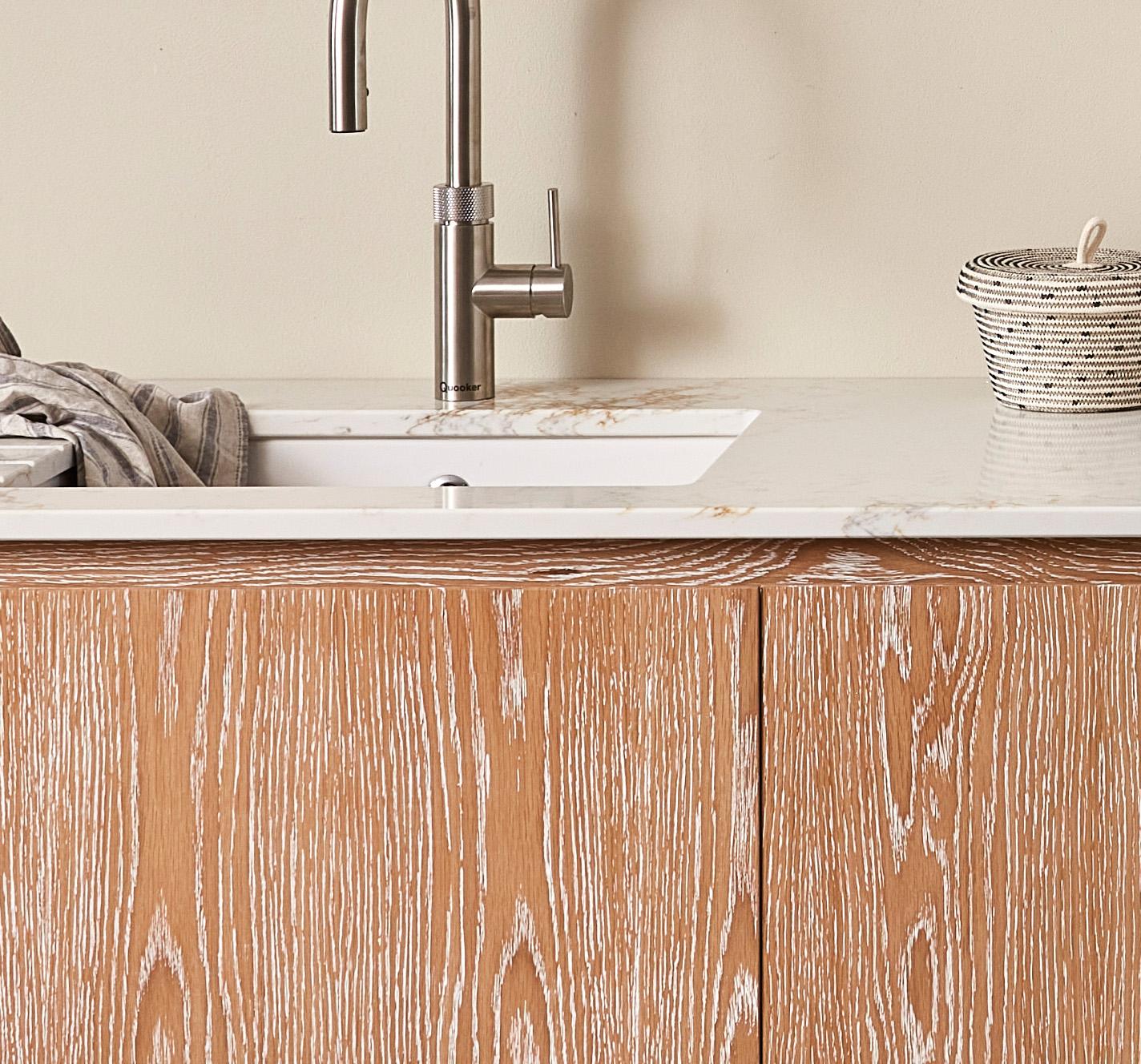 Wood-textured bathroom cabinet beneath a white countertop with a built-in sink. A sleek silver faucet is installed, with a gray towel draped on the side. A small, round, woven basket sits on the countertop against a light-colored wall.