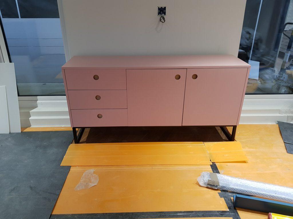 A modern pink sideboard with round cutout handles and black metal legs, set up in a room under construction.