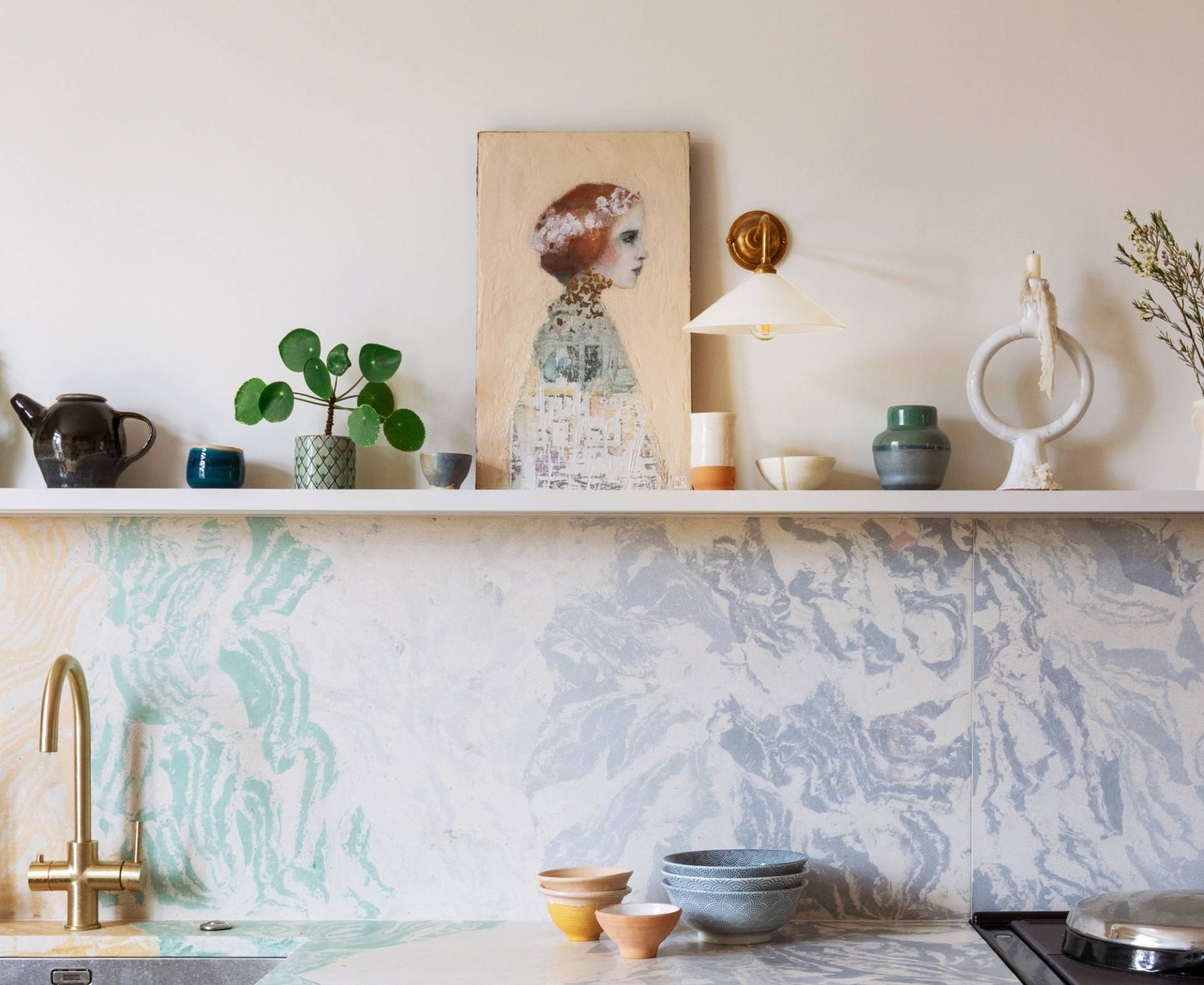 A modern kitchen with a marbled pastel backsplash, open shelf displaying pottery, plants, and artwork of a womans profile. Various bowls and cups are on the countertop beneath a brass faucet.