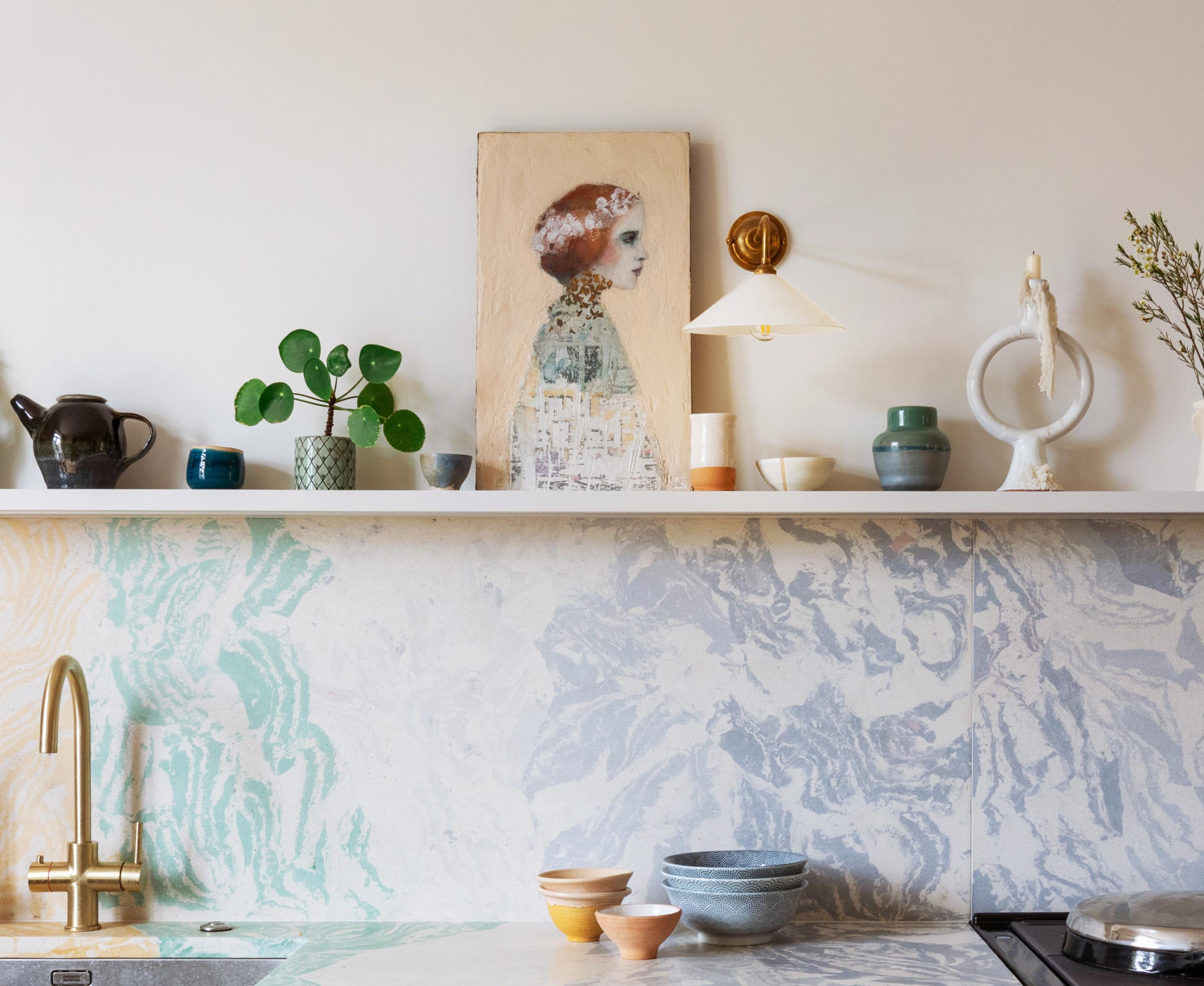A modern kitchen with a marbled pastel backsplash, open shelf displaying pottery, plants, and artwork of a womans profile. Various bowls and cups are on the countertop beneath a brass faucet.