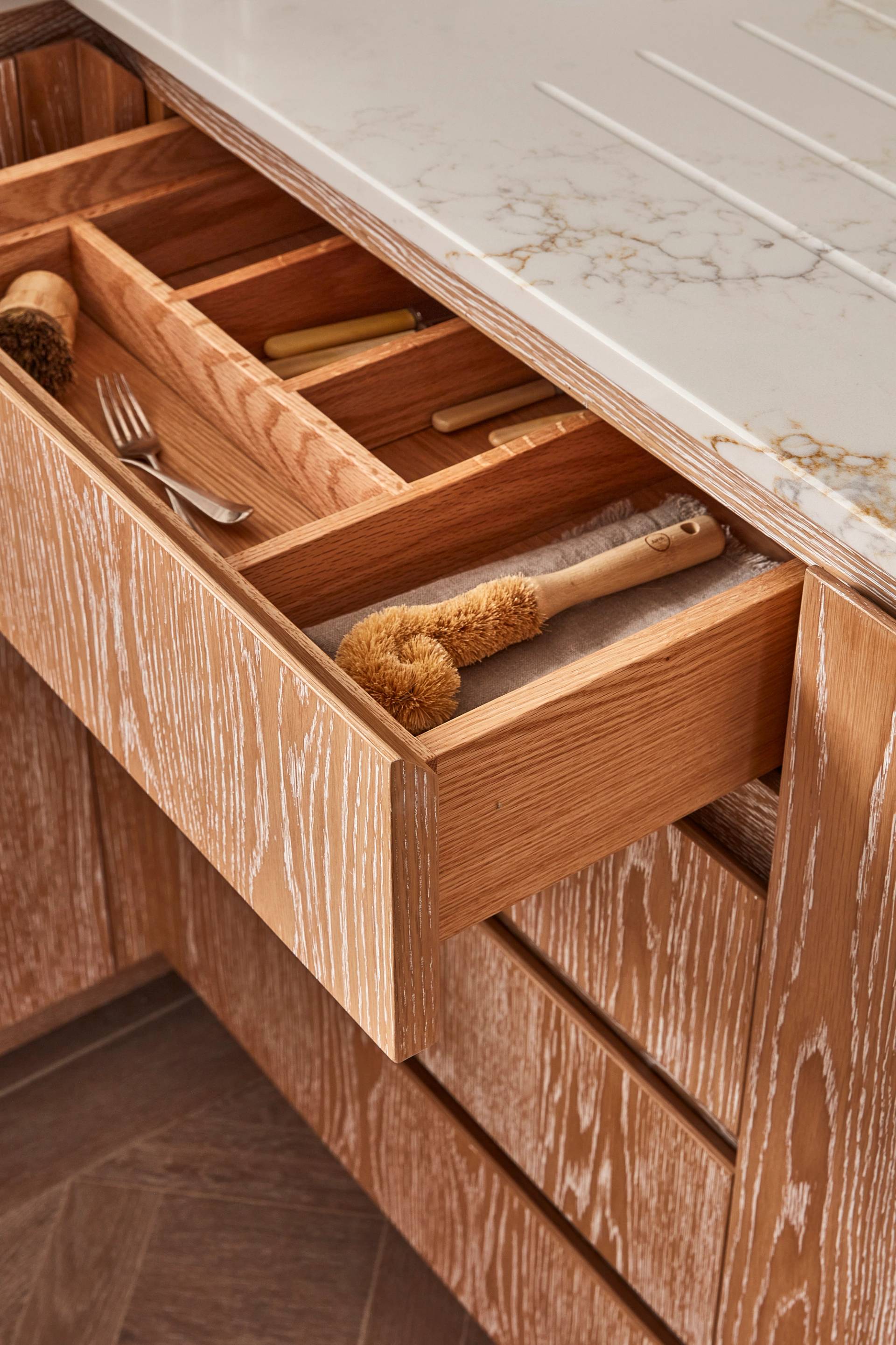 Open wooden kitchen drawer with divided compartments containing a scrub brush, fork, and other utensils. The drawer is beneath a light-colored countertop with subtle marble patterns, and the cabinet has a textured wood grain finish.