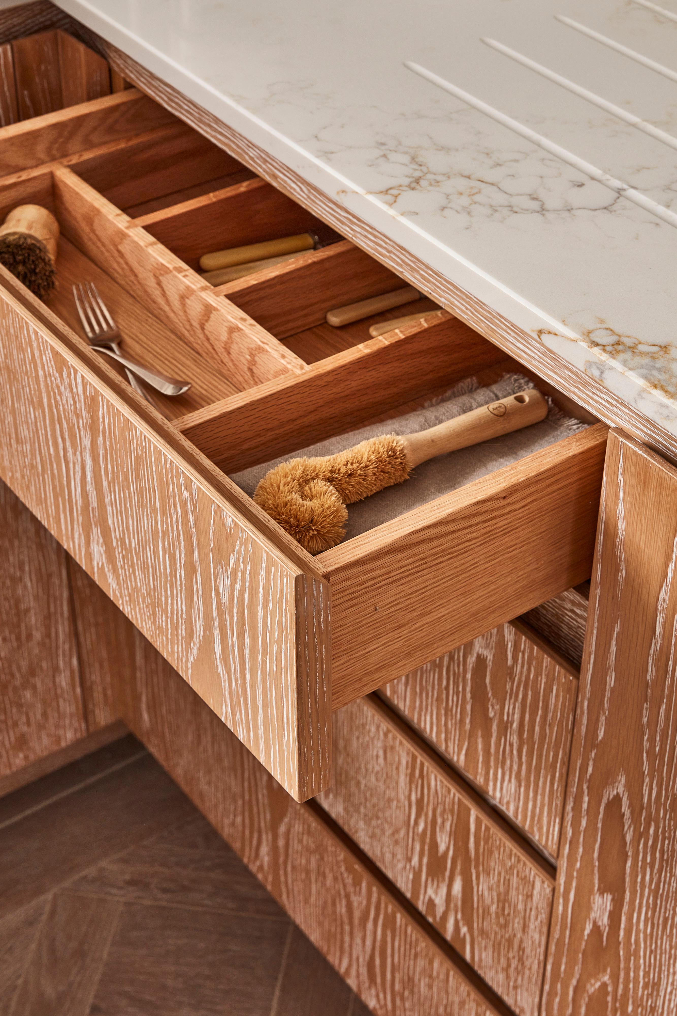 Open wooden kitchen drawer with divided compartments containing a scrub brush, fork, and other utensils. The drawer is beneath a light-colored countertop with subtle marble patterns, and the cabinet has a textured wood grain finish.