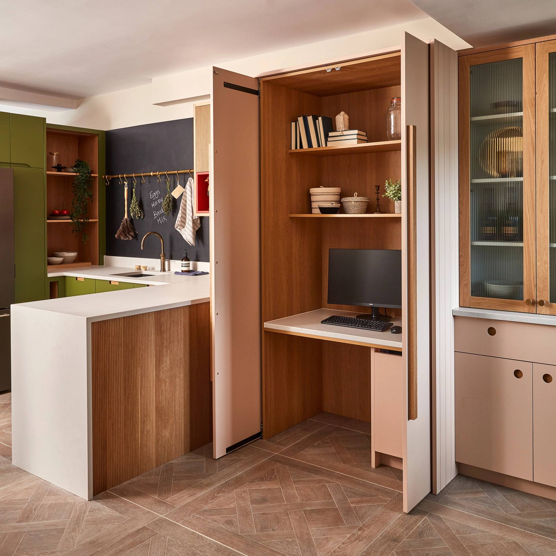 A modern kitchen with wooden cabinetry and green accents. A sliding door reveals a compact home office nook with a desk, computer, and chair. The space features an island with a white countertop and recessed lighting, with potted plants for decor.