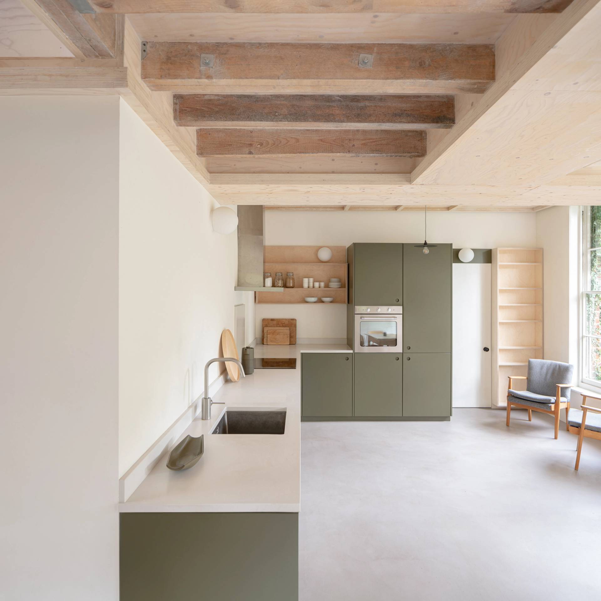 Minimalist kitchen with light green cabinets, white countertops, and open wooden shelves. Ceiling features exposed wooden beams. A chair and a small table are placed near a window, allowing natural light to brighten the space.