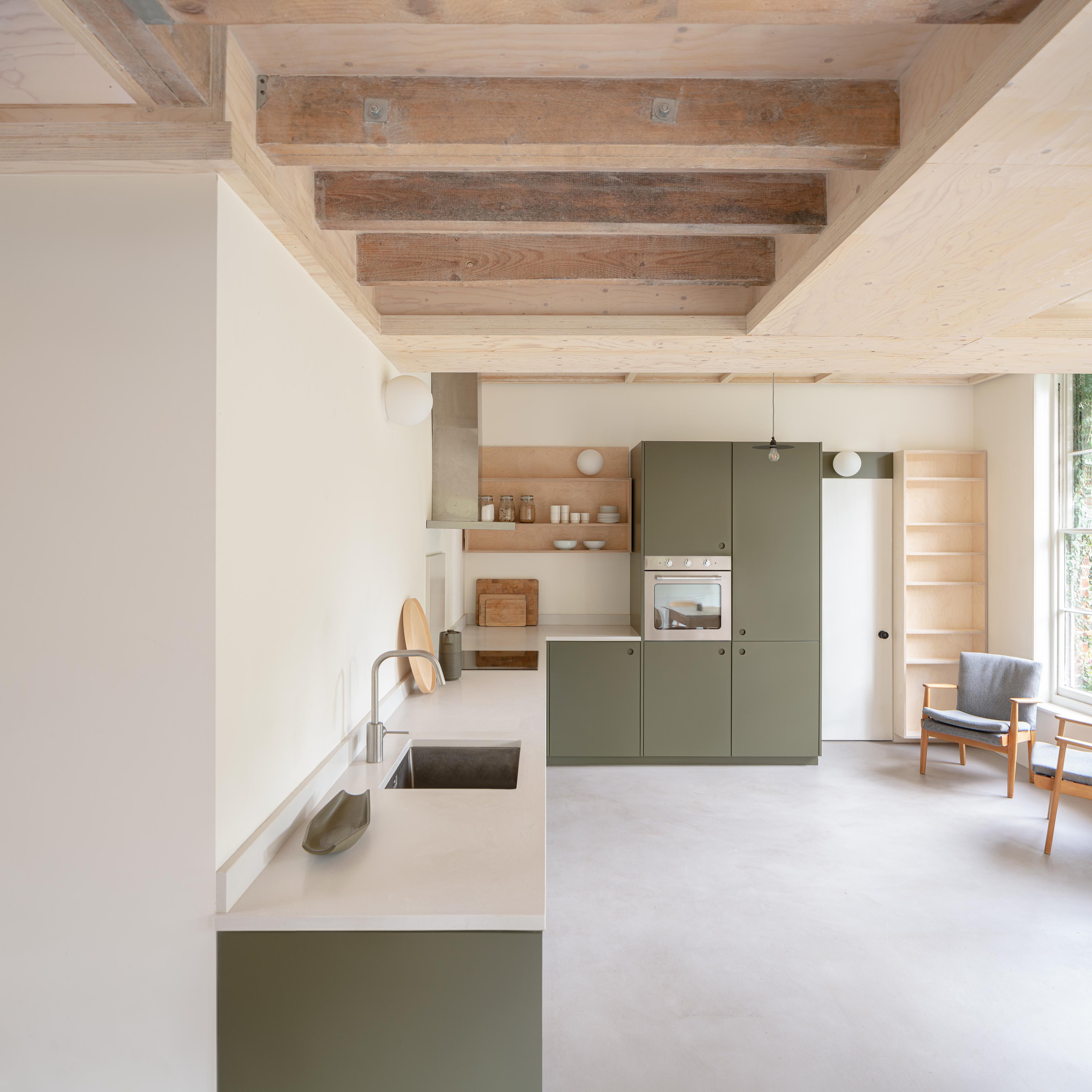 Minimalist kitchen with light green cabinets, white countertops, and open wooden shelves. Ceiling features exposed wooden beams. A chair and a small table are placed near a window, allowing natural light to brighten the space.