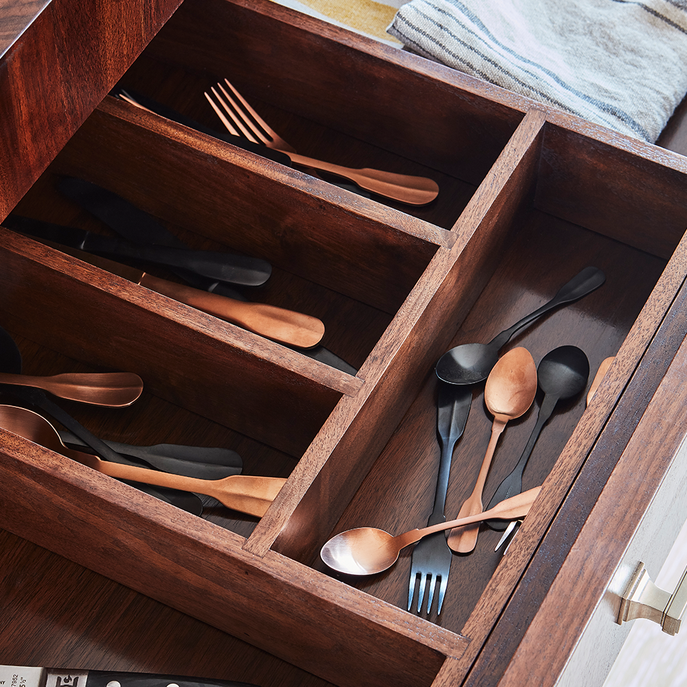 A wooden cutlery drawer is organized into sections, containing black and copper-colored forks and spoons, some stacked and some scattered, with a folded towel visible in the top corner.