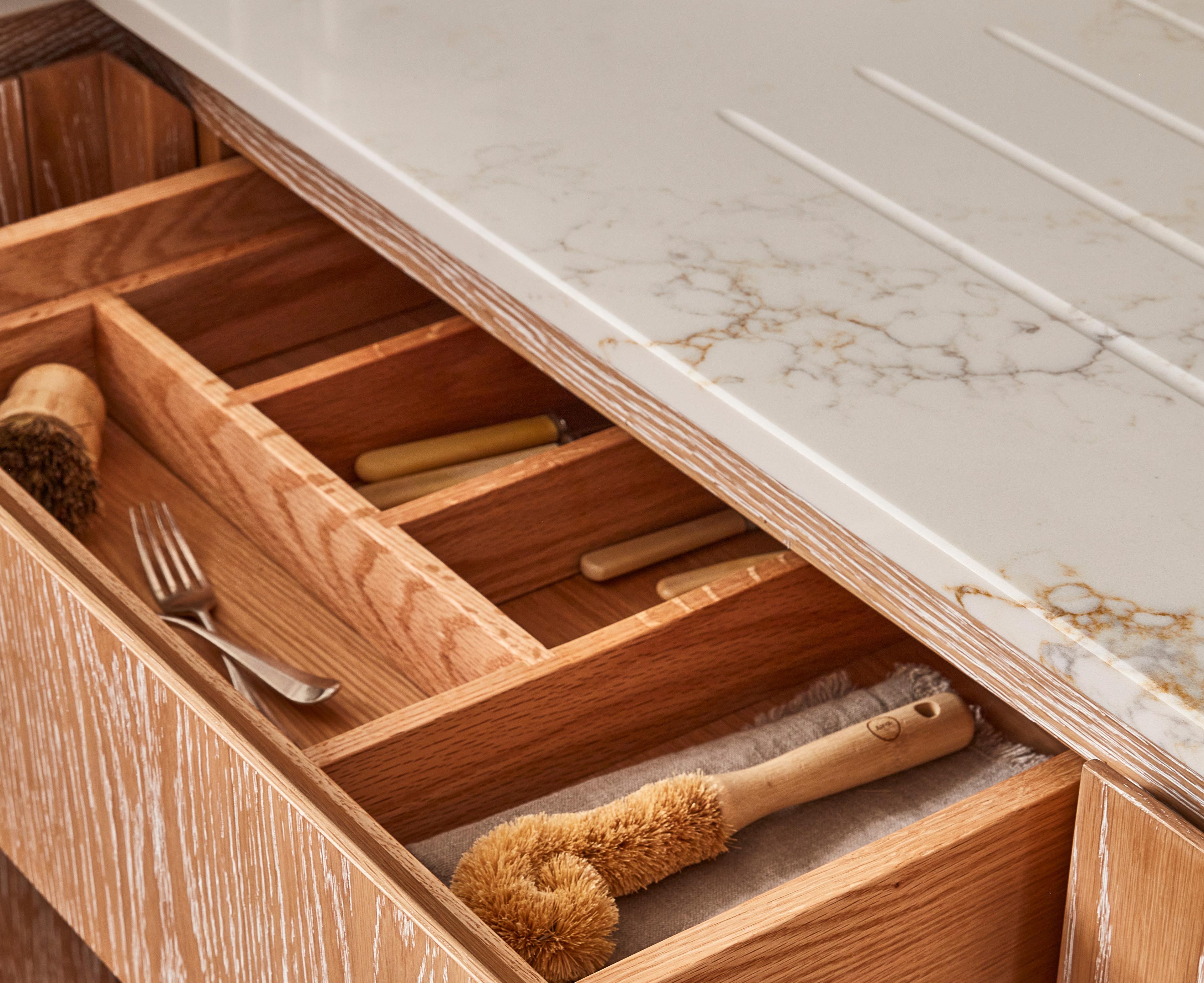 Open wooden kitchen drawer with organized sections containing wooden utensils and a brush. The countertop above is white marble with subtle gray veining.