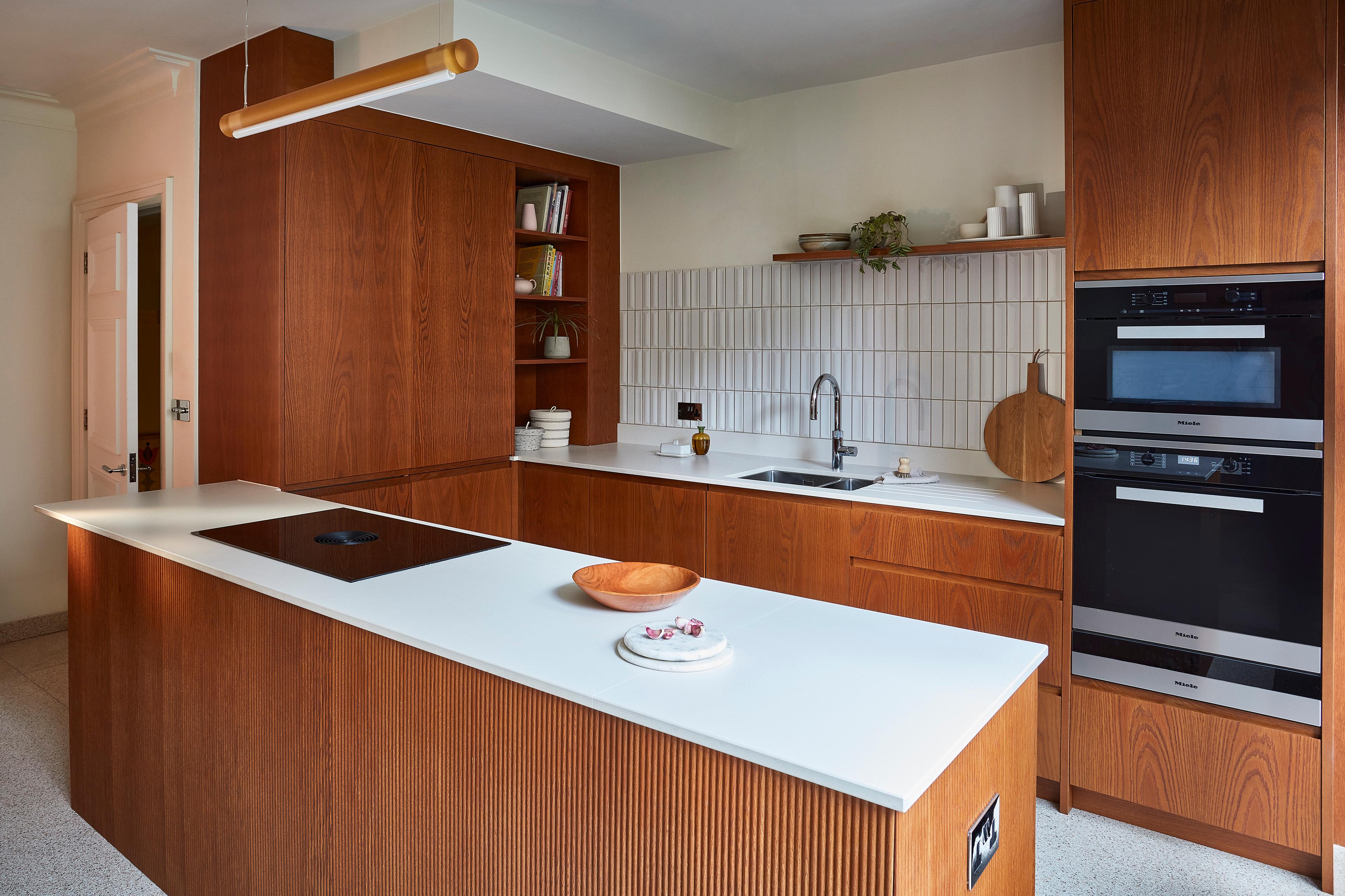 Modern kitchen with wooden cabinets, white countertops, built-in black ovens, a cooktop, open shelving with dishes, a sink, and a tiled backsplash. A wooden bowl and a plate with treats are on the island.