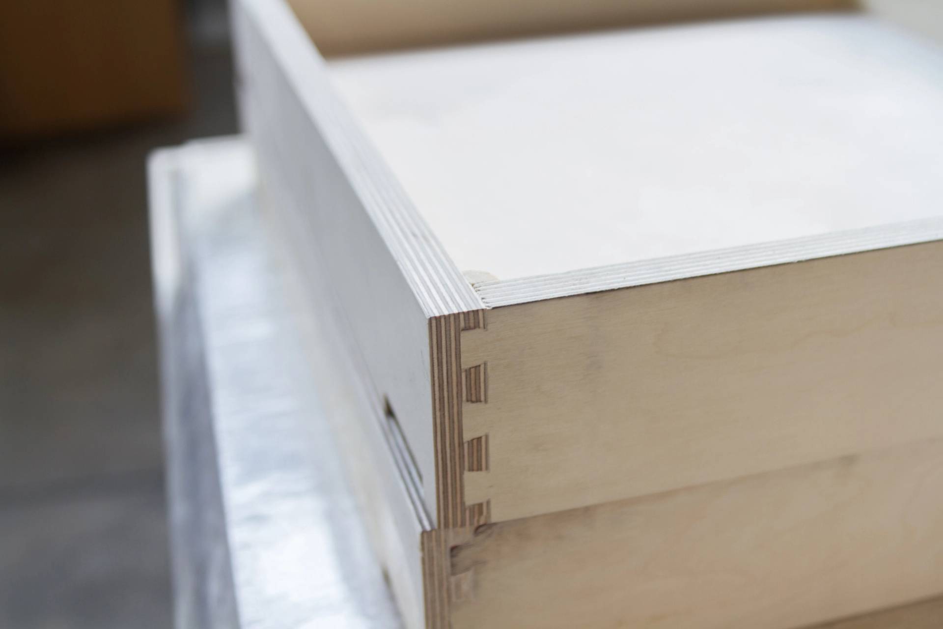 Close-up of a wooden box corner showing interlocking dovetail joints. The wood grain is visible, and the box is positioned on a reflective surface in a workshop setting.