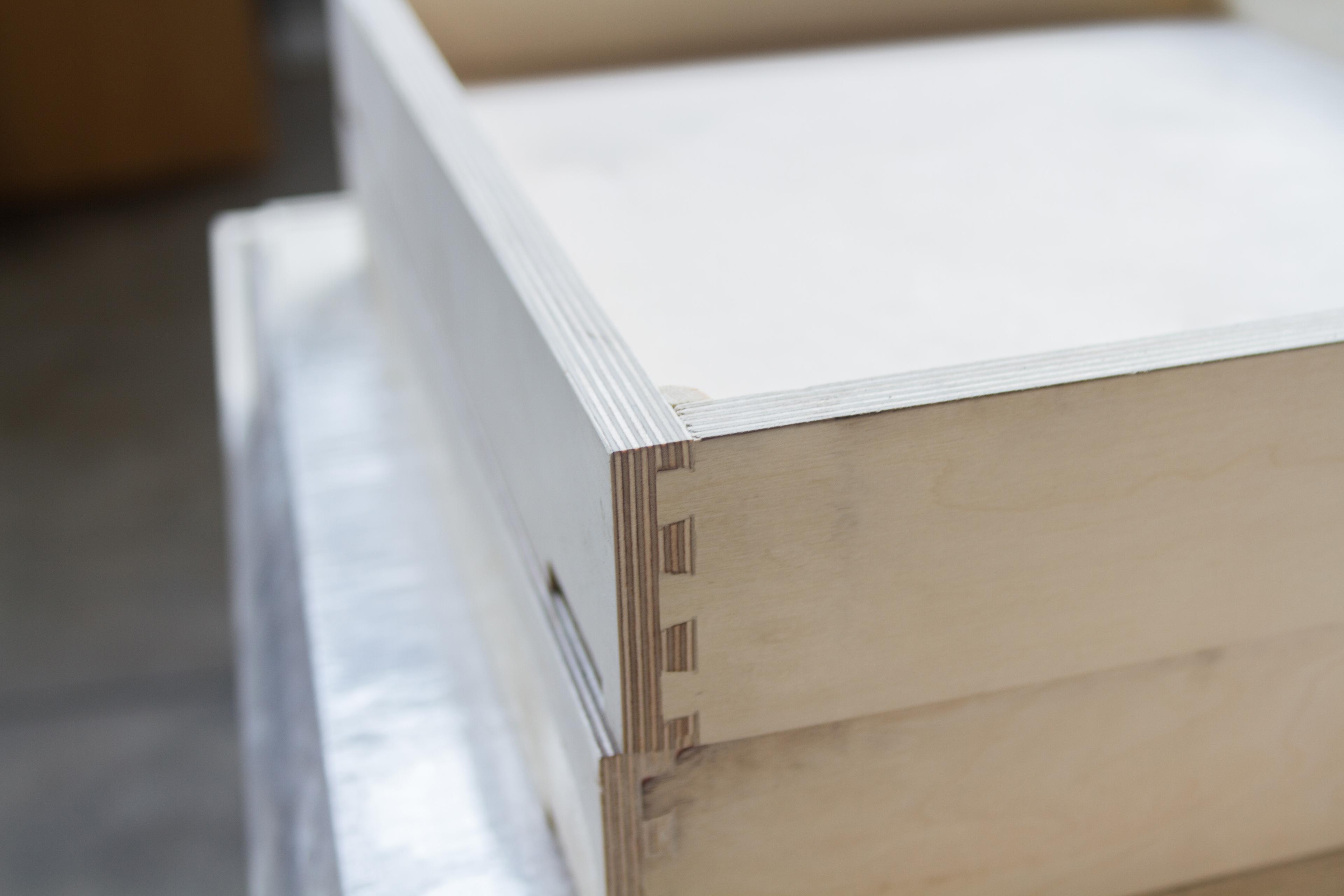 Close-up of a wooden box corner showing interlocking dovetail joints. The wood grain is visible, and the box is positioned on a reflective surface in a workshop setting.