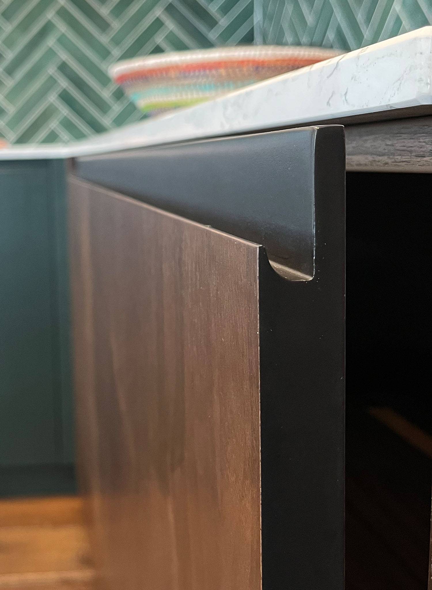 Close-up view of a modern kitchen cabinet with a matte black handle integrated into the top edge of a dark wood door, beneath a white countertop and green tile backsplash.