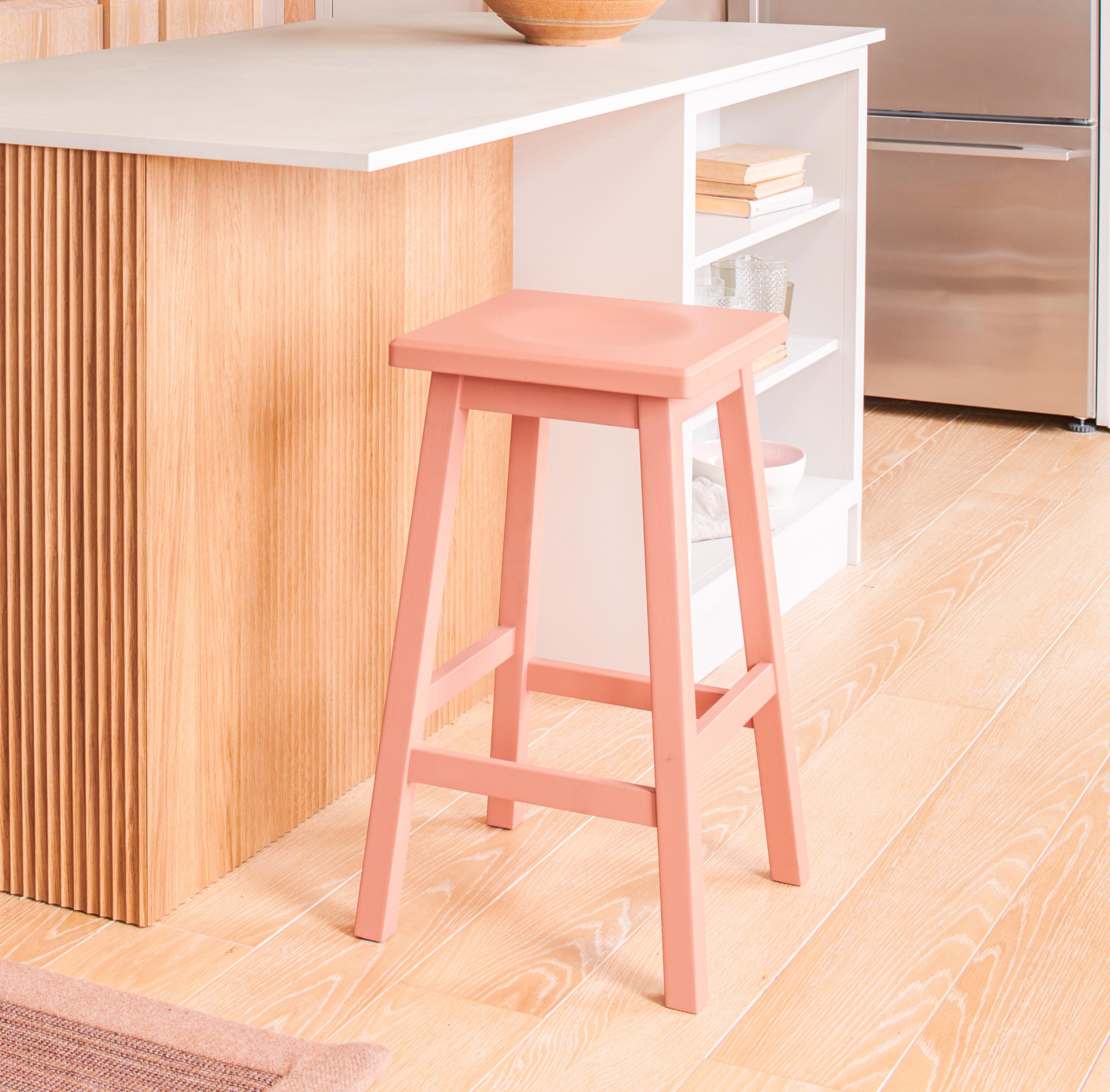 A modern kitchen with light wood flooring features a salmon pink wooden stool next to a beige island with a wood paneling accent. The island has white shelving and a silver refrigerator is visible in the background.