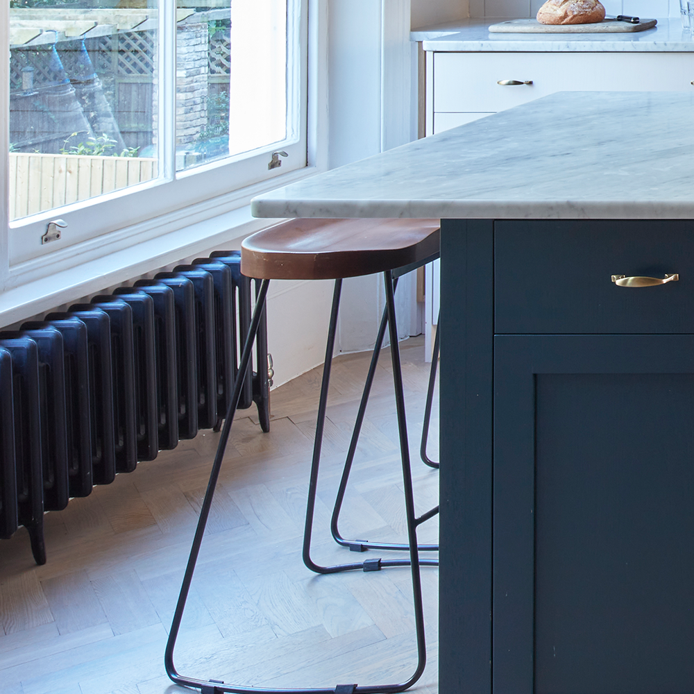 A modern kitchen interior with a marble countertop island, a wooden barstool with metal legs, a radiator under a large window, and light wood herringbone flooring.
