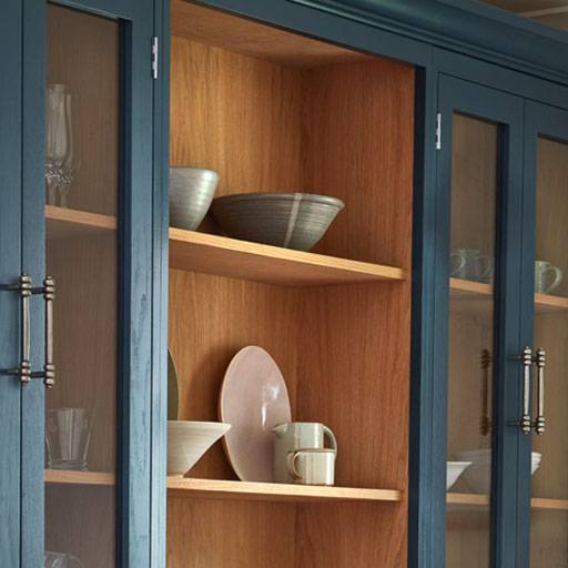 A wooden kitchen cabinet with glass doors displays ceramic bowls, plates, cups, and glasses on its shelves. The outer frame is painted dark blue, while the interior has a natural wood finish.