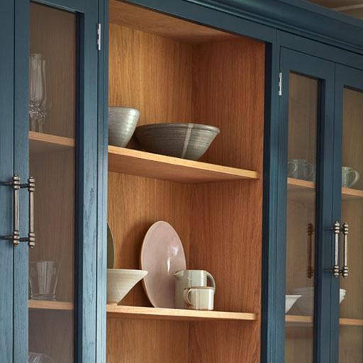 A wooden kitchen cabinet with glass doors displays ceramic bowls, plates, cups, and glasses on its shelves. The outer frame is painted dark blue, while the interior has a natural wood finish.