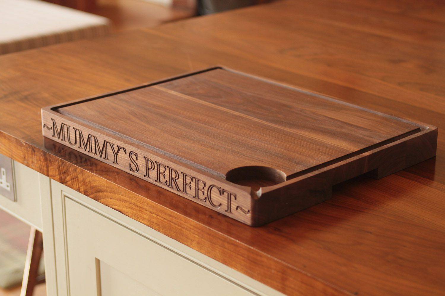 Wooden chopping board on a kitchen counter, featuring carved text that reads 'mummys perfect'
